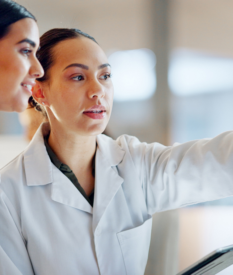 Two female researchers in lab coats analyzing data on a computer in a laboratory, with a microscope in the foreground – illustrating digital transformation and data-driven work in the life sciences sector.